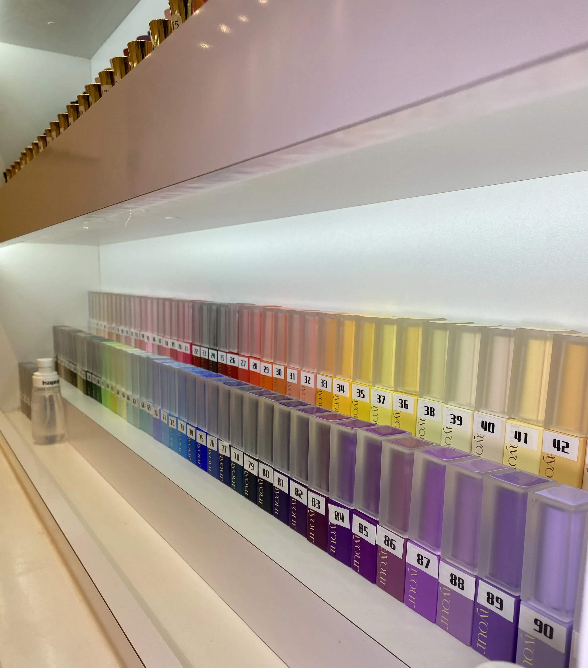 Display shelf with rows of colorful nail polish bottles labeled with numbers, arranged in a gradient from pink to purple, with a small bottle of hand sanitizer nearby.