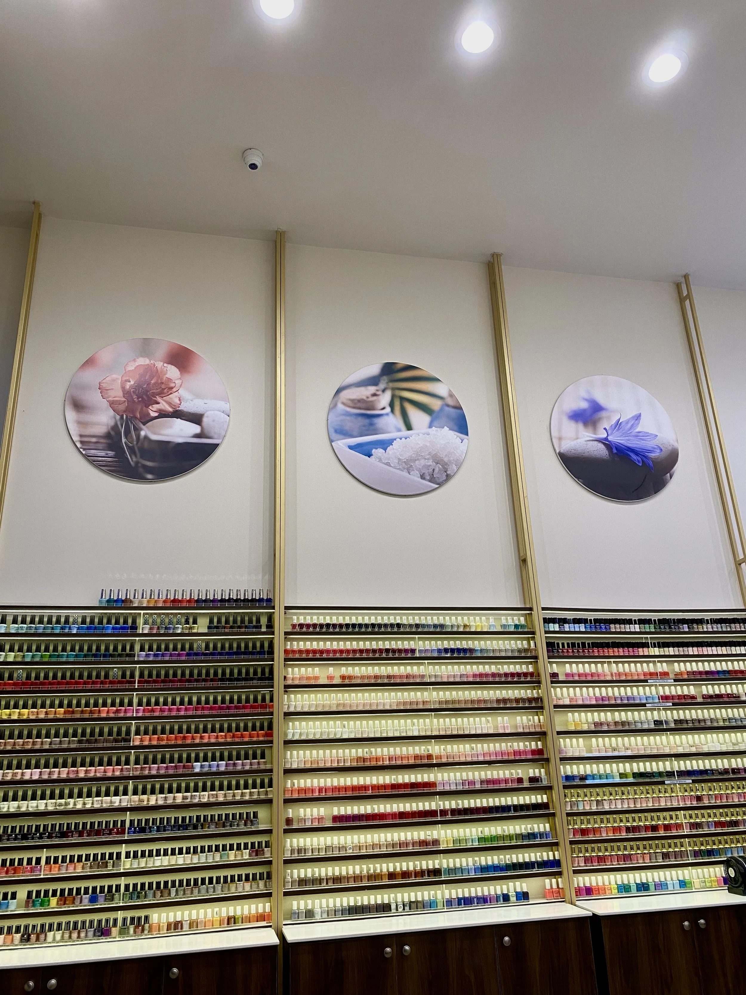 Wall display of nail polish bottles arranged on shelves below three circular art prints of spa or massage scenes.