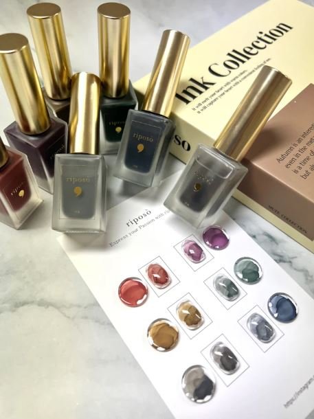 Nail polish bottles with gold caps, a color swatch sheet with various shades, and a box labeled 'Ink Collection' on a marble surface.