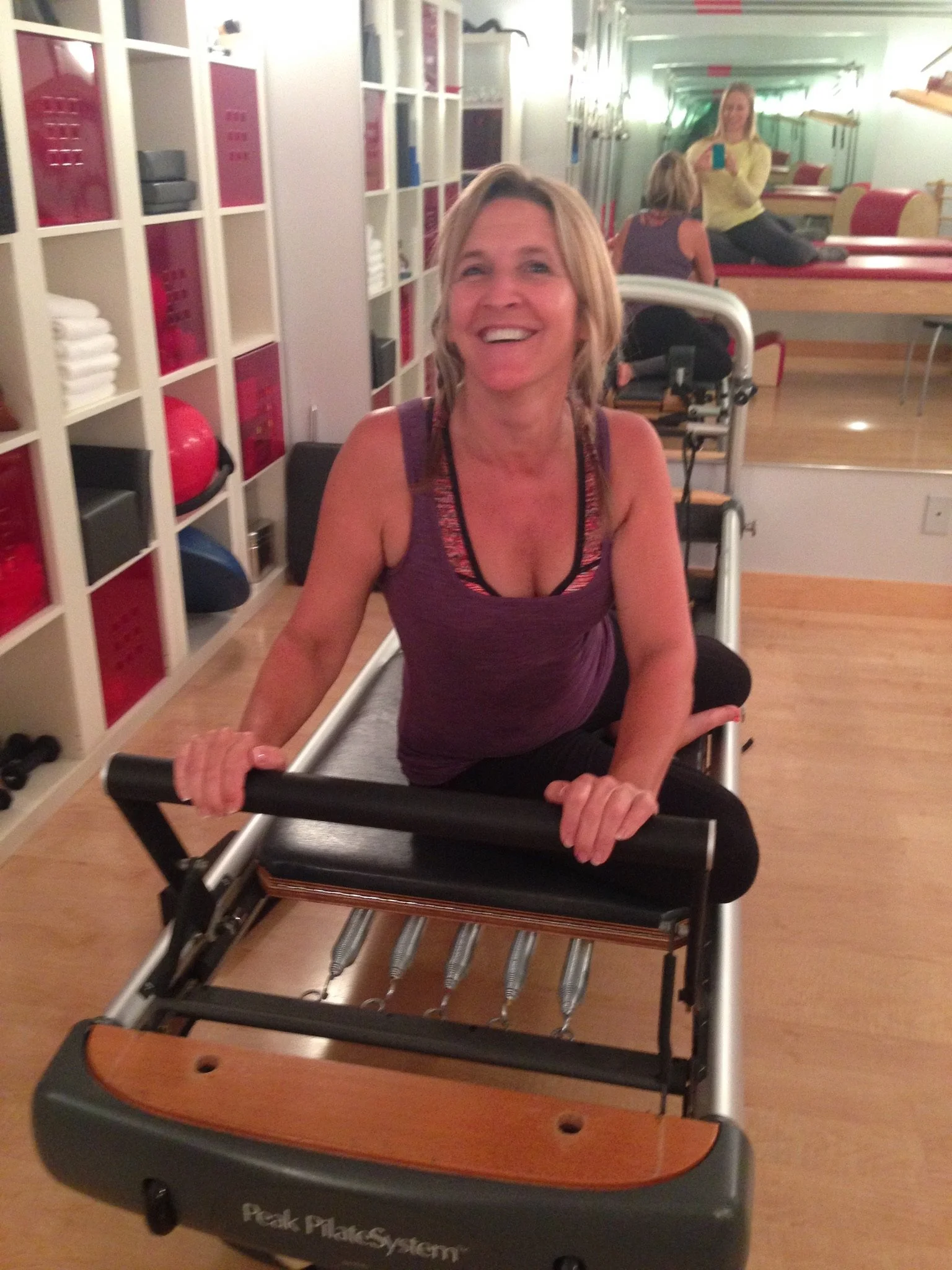 Woman is sitting on the pilates reformer machine doing a pilates exercise. She is smiling while working hard and feeling the burn.