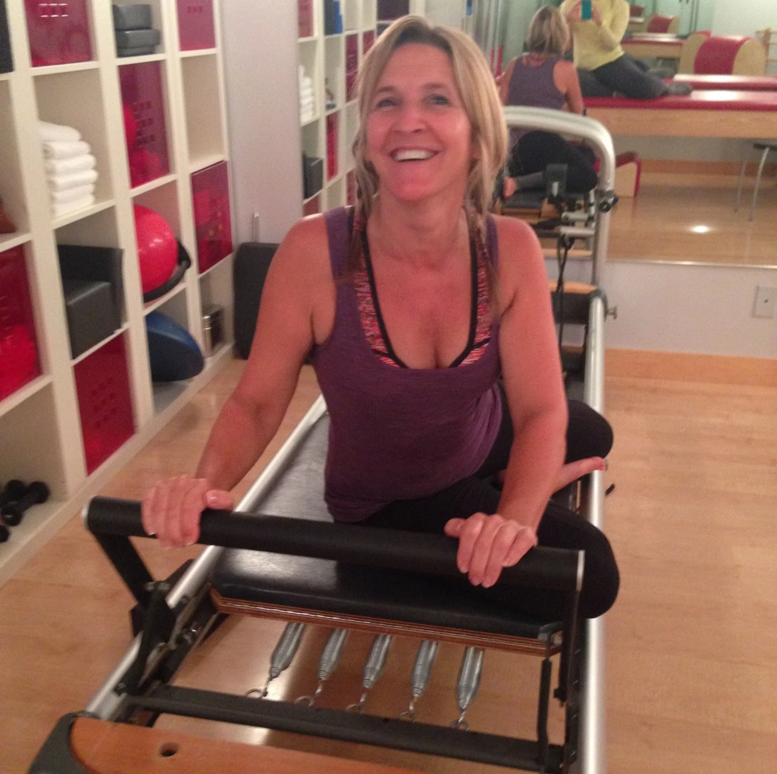 We love pilates! She is on a pilates reformer and having a private lesson to learn the ins and outs of pilates fitness. The reformer has springs and a bar that she is holding on to. She is sitting on her side doing a exercise while twisting.