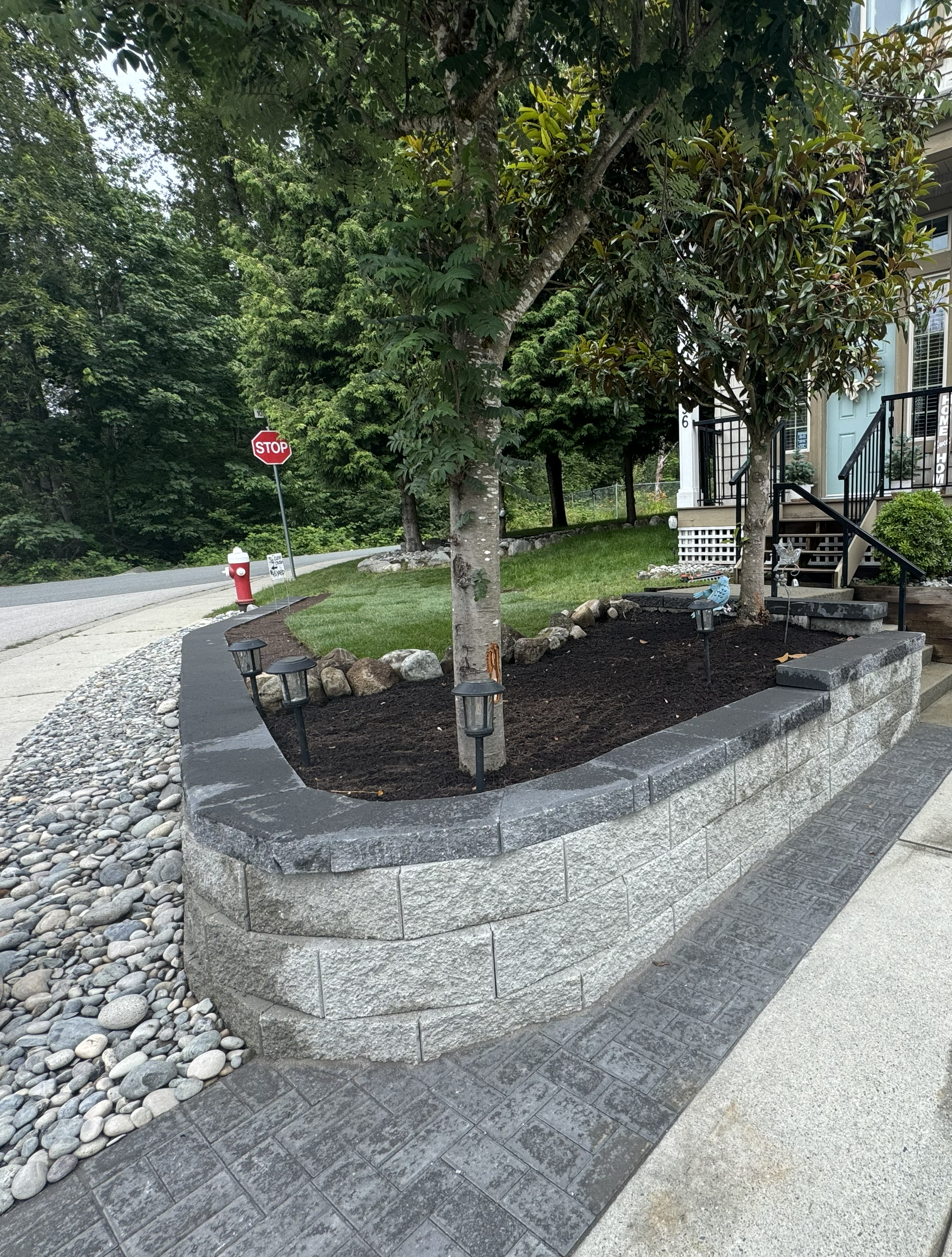 Front yard with a tree on the left, surrounded by rocks and small bushes, leading to a house with gray garage doors, stairs with wooden railings, and an open porch above.