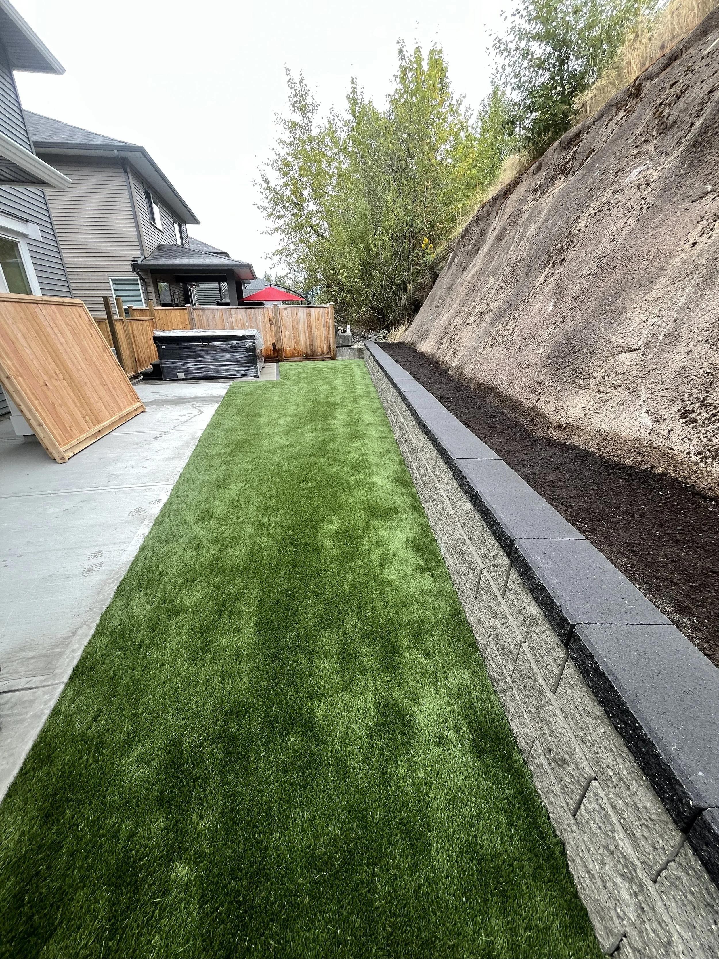 Backyard with artificial grass lawn, wooden fence, and retaining wall next to a steep dirt hillside. There are some outdoor furniture pieces and a hot tub near the fence.