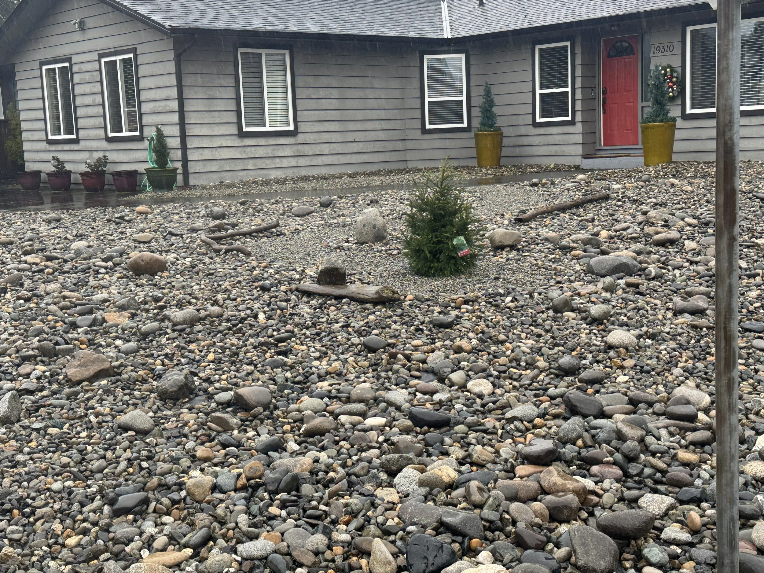 Front yard with a tree on the left, surrounded by rocks and small bushes, leading to a house with gray garage doors, stairs with wooden railings, and an open porch above.