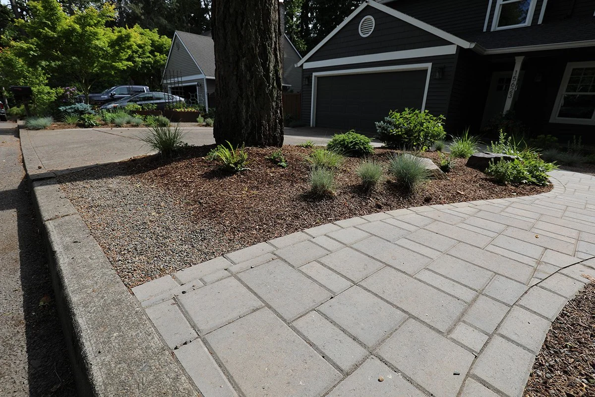 Residential driveway and front yard with landscaped garden, tree, and black garage door.
