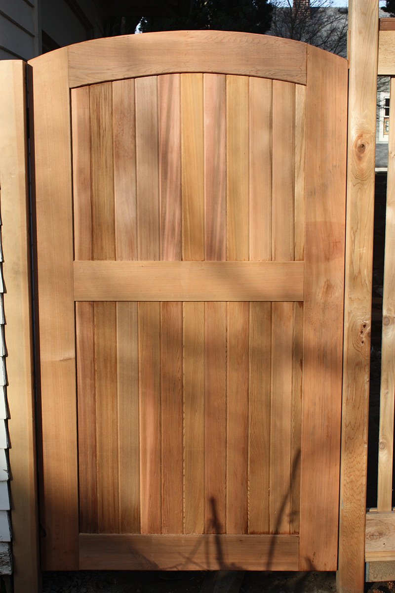 Unpainted wooden gate with vertical planks and an arched top, installed outdoors with part of a wooden fence and siding visible on the sides.