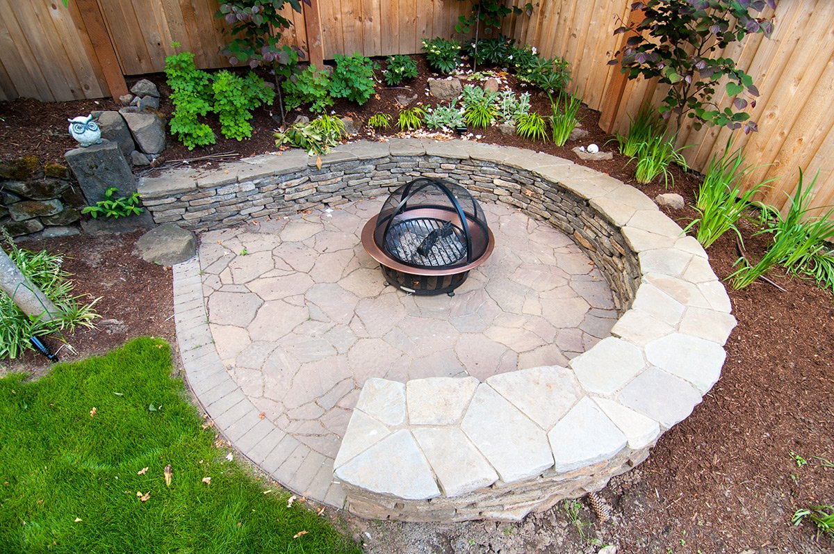 A backyard patio with a circular stone fire pit, surrounded by a stone border, and a garden with various green plants and a wooden fence in the background.