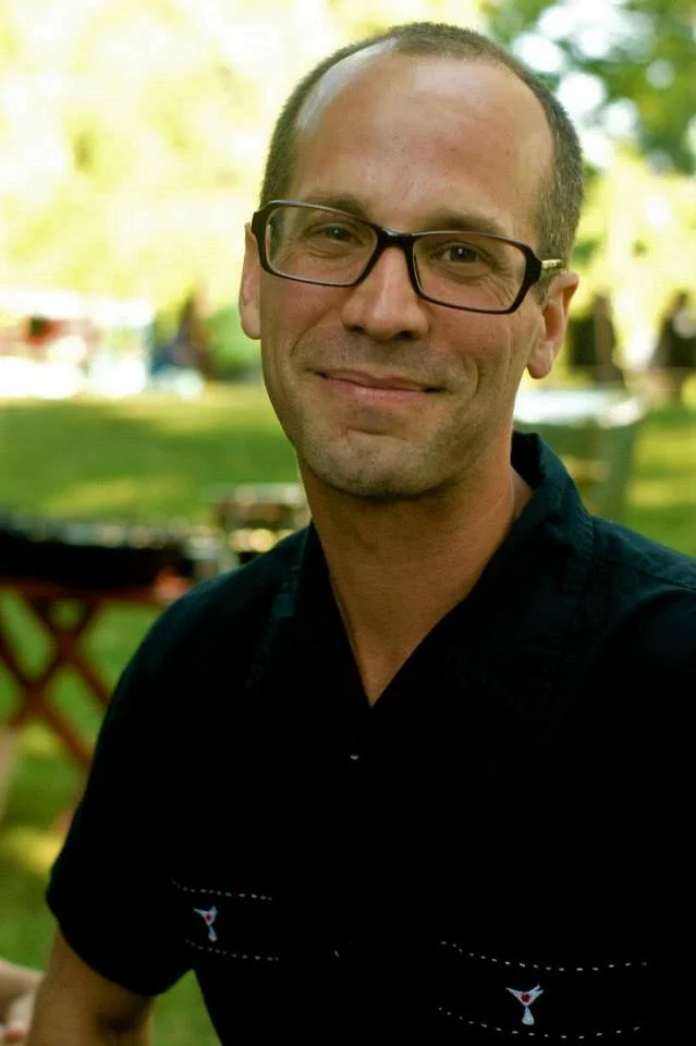 Man with glasses standing outdoors in a park or garden, smiling at the camera.