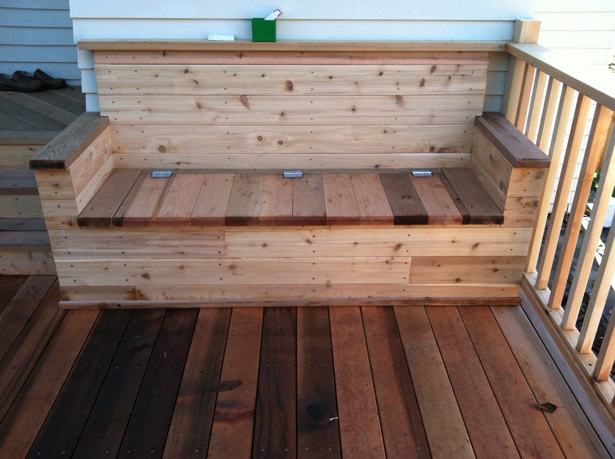 Wooden deck with a built-in wooden bench, partially stained with darker wood accents, located outside a house with white siding and a wooden railing.