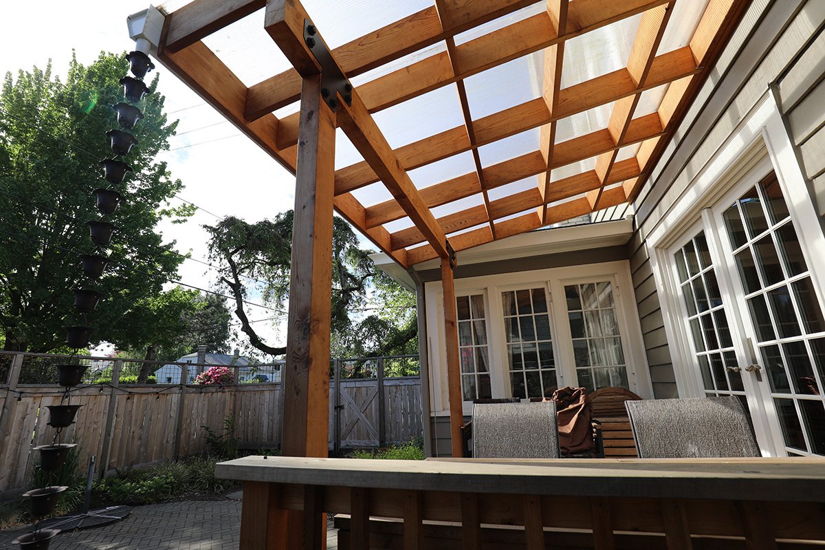Back porch with wooden pergola under construction, patio furniture, potted plants, and a wooden fence in the background.
