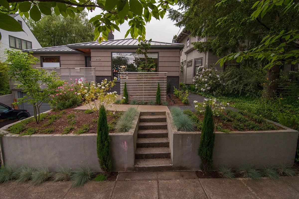 Front yard with elevated garden beds, small steps, and a modern house with large windows, surrounded by trees and plants.