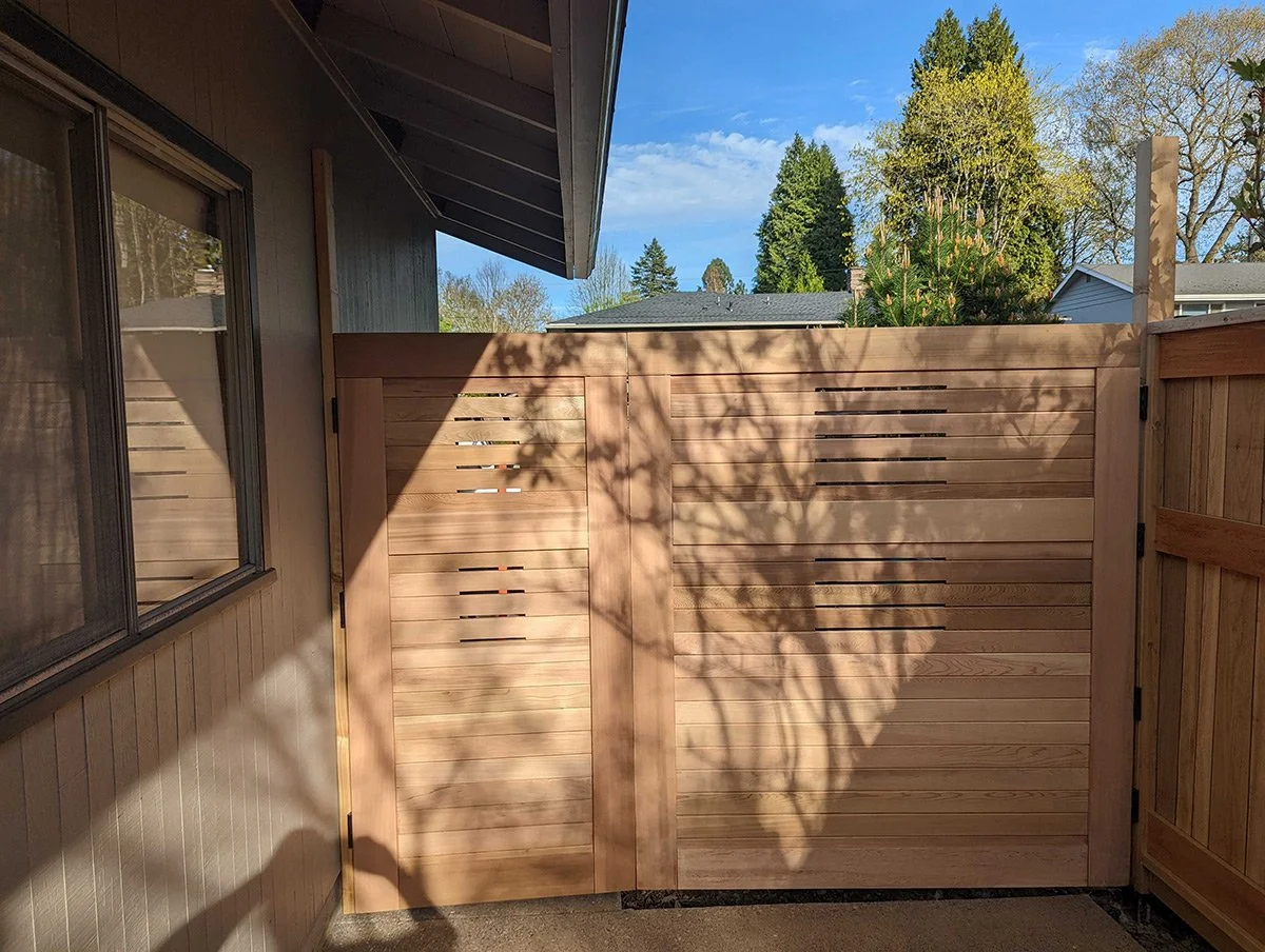 Wooden privacy fence with shadow of tree branches, part of house exterior and window in a backyard during daytime.