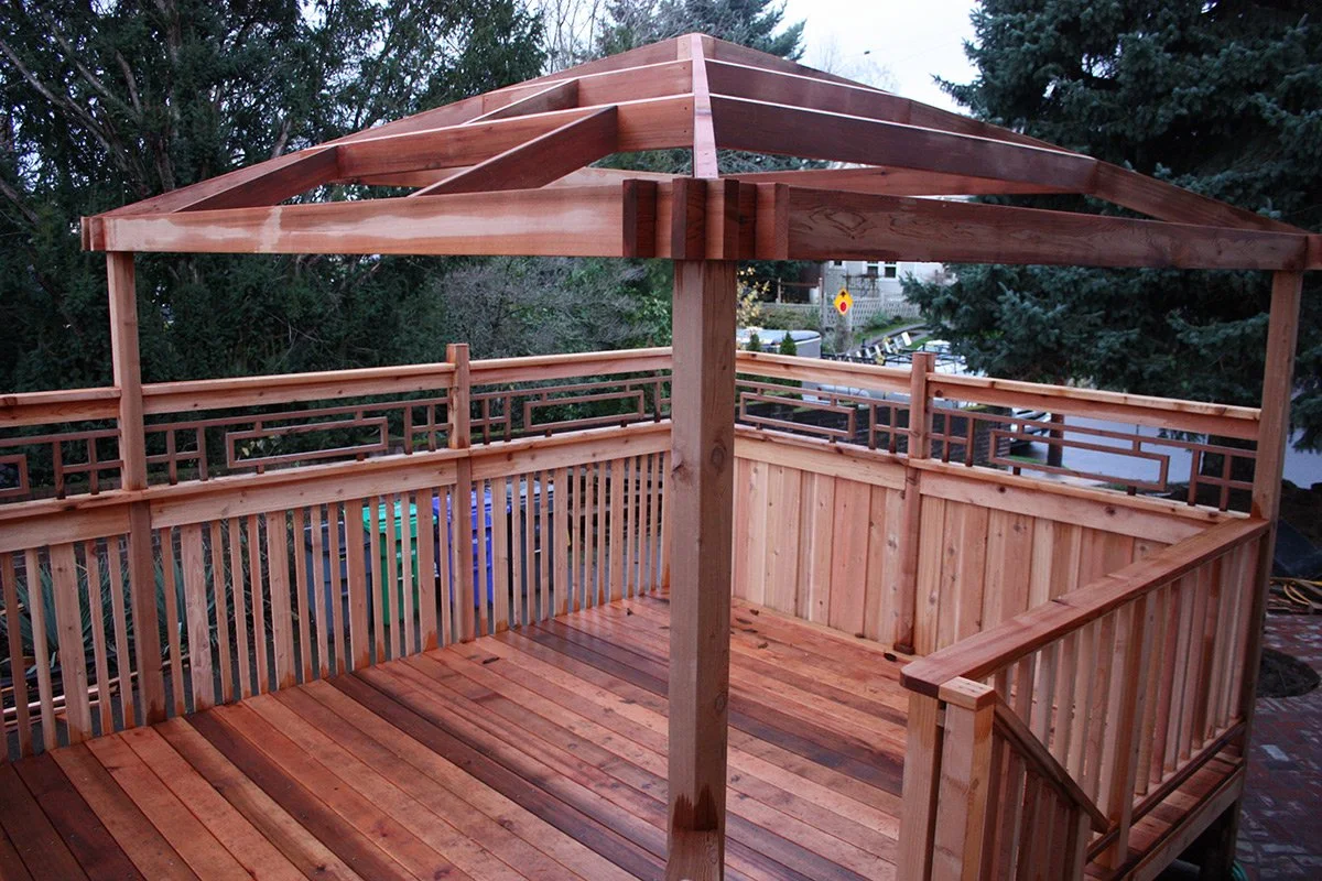 A wooden deck with a partially constructed pergola frame, featuring vertical posts, angled support beams, and decorative railing, surrounded by trees and neighboring houses.