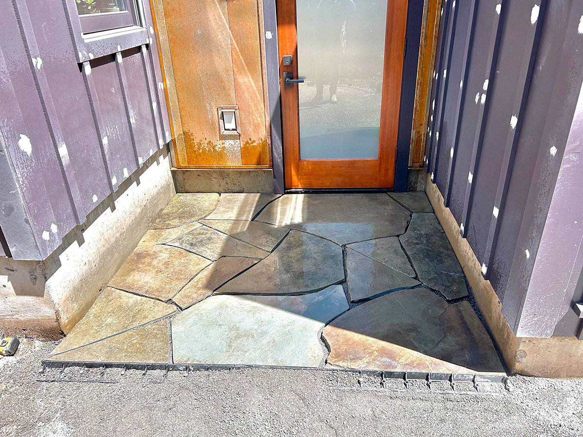 Slate stone walkway in front of a wooden door, with purple wooden paneling on either side and a concrete border.