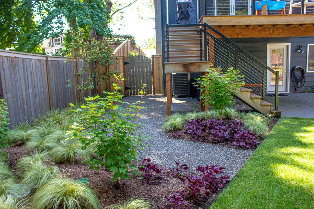 Backyard garden with gravel pathway, plants, and shrubs, wooden fence, deck stairs with metal railing, house exterior with door, window, and garden hose