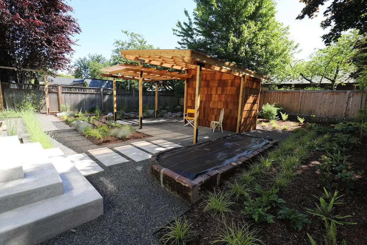 A backyard with a garden, stone pathway, small wooden shed with overhang, a chair, and plants, surrounded by a wooden fence.