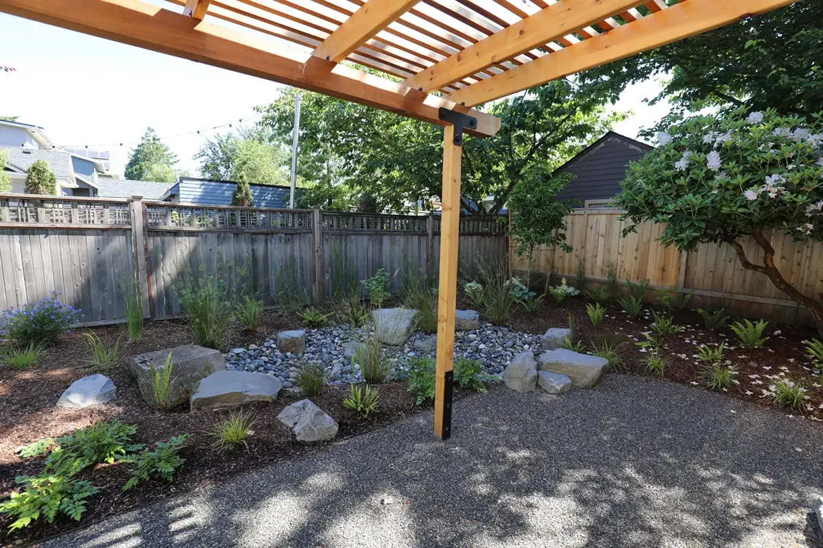 Backyard garden with wooden pergola, landscaping rocks, and flowering plants, enclosed by wooden fence.