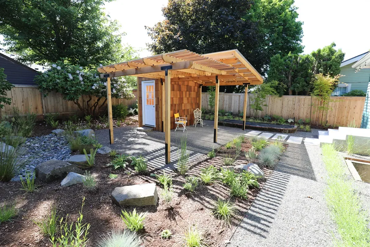 A backyard garden with a wooden structure with a slanted roof, a white door, and outdoor chairs. There are plants and flowers in the garden beds and a stone pathway, surrounded by a wooden fence with trees in the background.