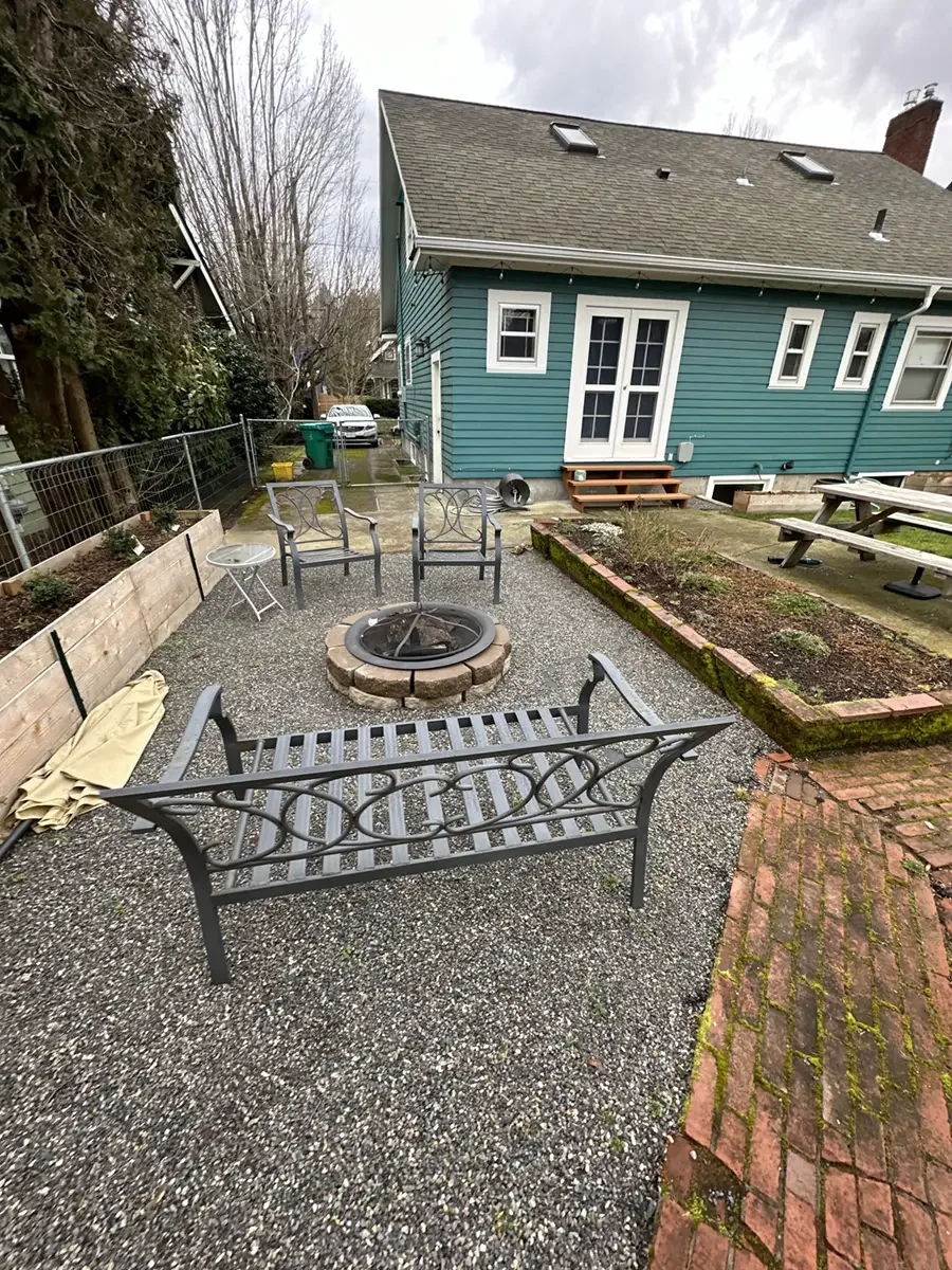 Backyard patio with metal benches arranged around a fire pit, brick and gravel flooring, and a raised garden bed with plants. A blue house with several windows and a door is in the background. There are some trees and a fence along the side.