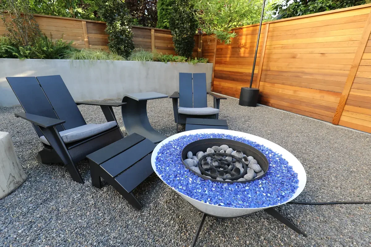 Outdoor backyard patio with modern fire pit surrounded by four black Adirondack chairs and two modern side tables, enclosed by wooden fence and greenery.