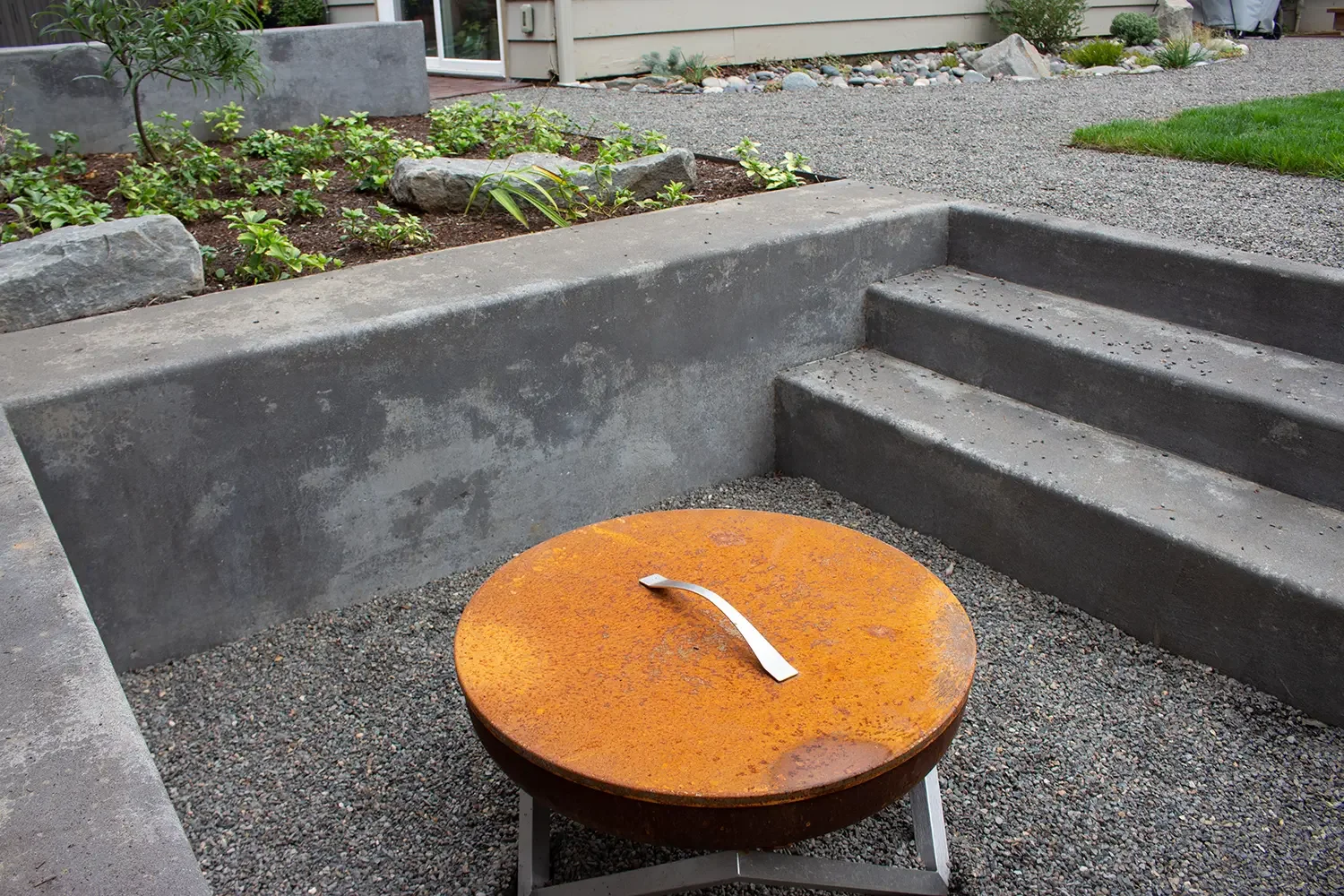 A rusted round fire pit with a metal handle on top, situated on gravel ground in a backyard with concrete steps and a garden bed with plants and rocks.
