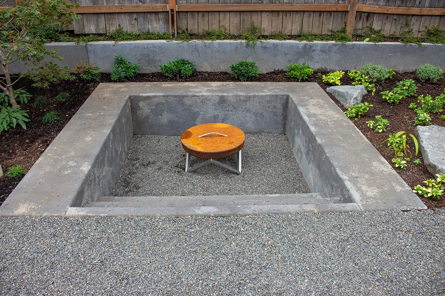 Concrete fire pit with a rusted metal cover, surrounded by gravel, in a backyard garden with plants and a wooden fence.