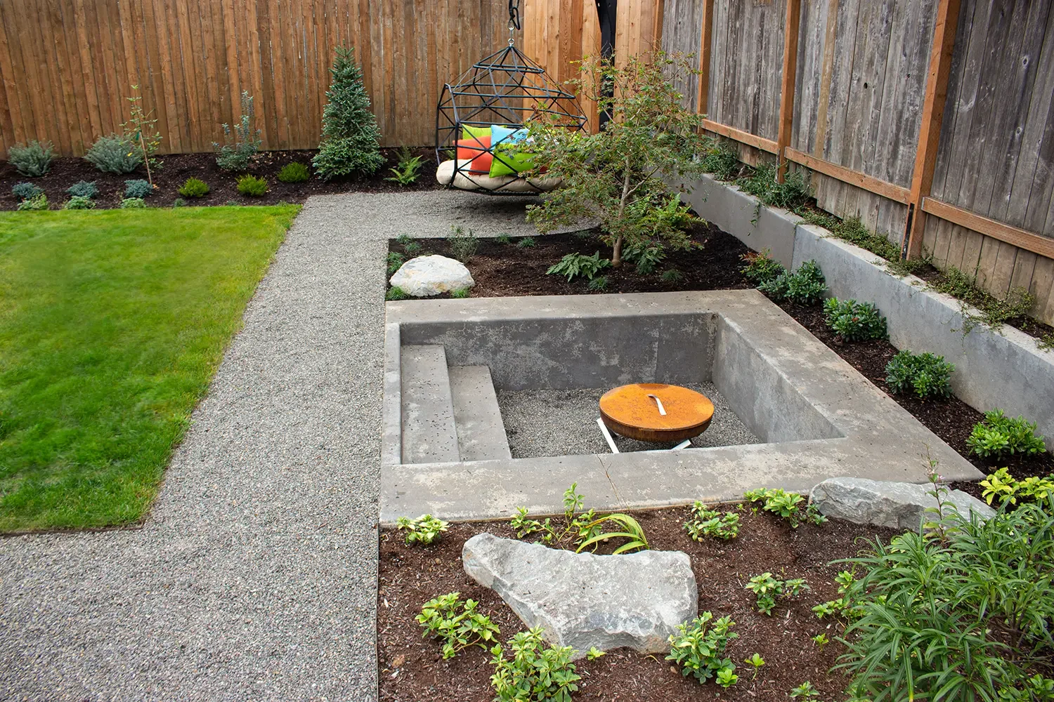 Backyard garden with a small grassy area, gravel pathway, and a fire pit area surrounded by rocks and plants. A seating area with colorful cushions is on a hanging chair near the back fence.
