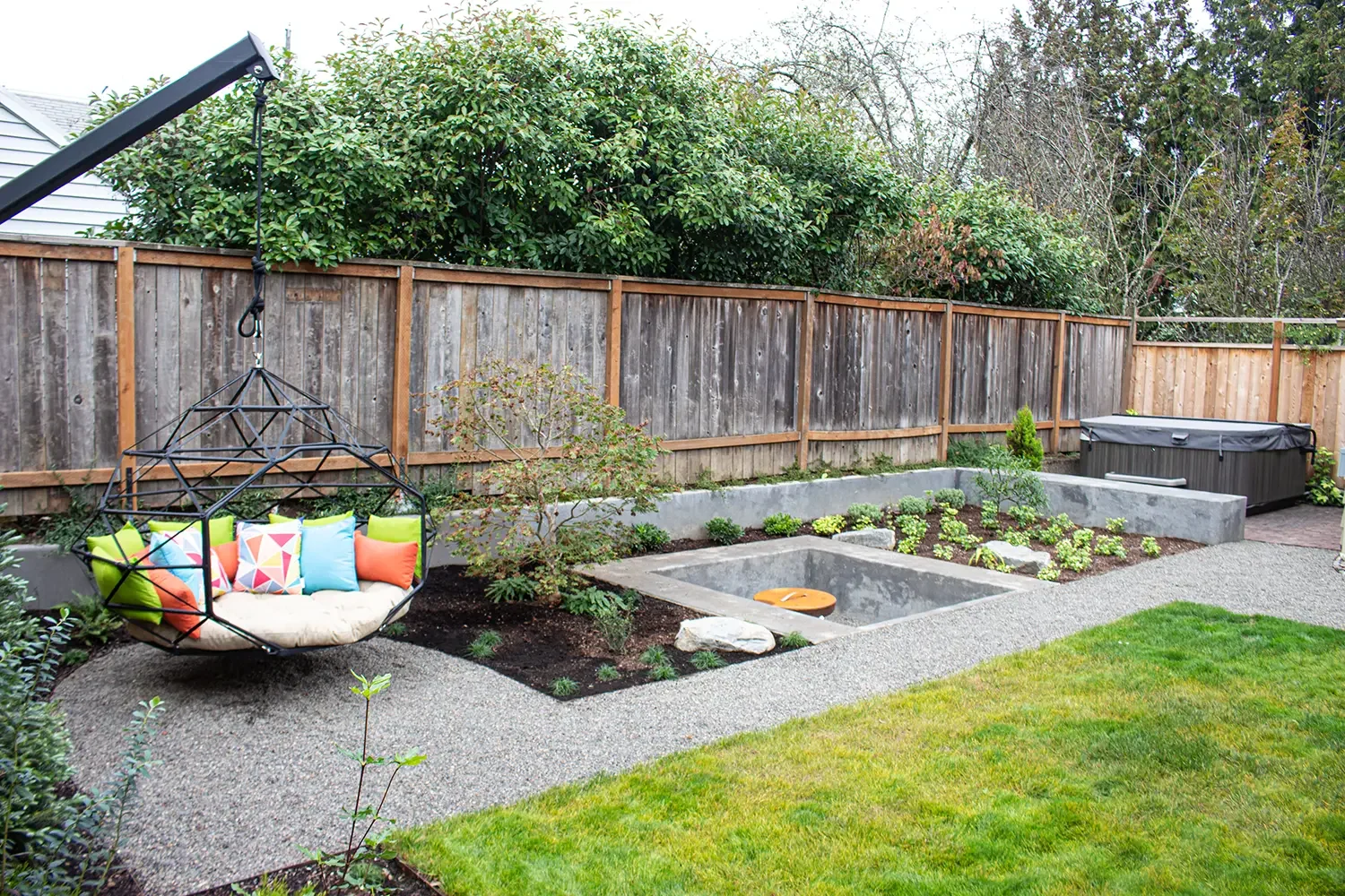 A backyard garden with a wooden fence, a hanging circular swing with colorful pillows, small landscaped plants, and a hot tub in the corner.