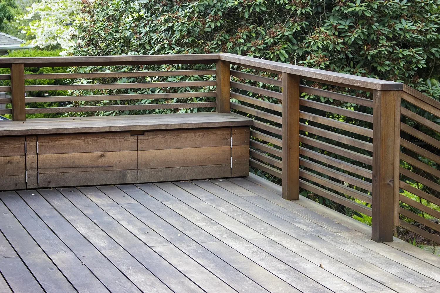 Wooden patio deck with a built-in wooden storage box and a railing, surrounded by greenery and bushes.