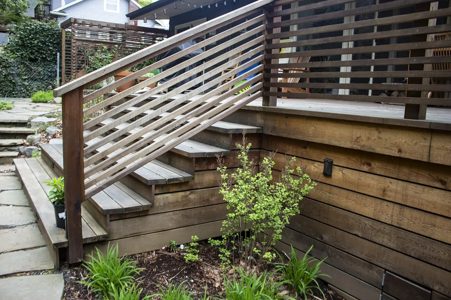 Wooden deck with stairs, railing, and small plants and bushes in a backyard garden.