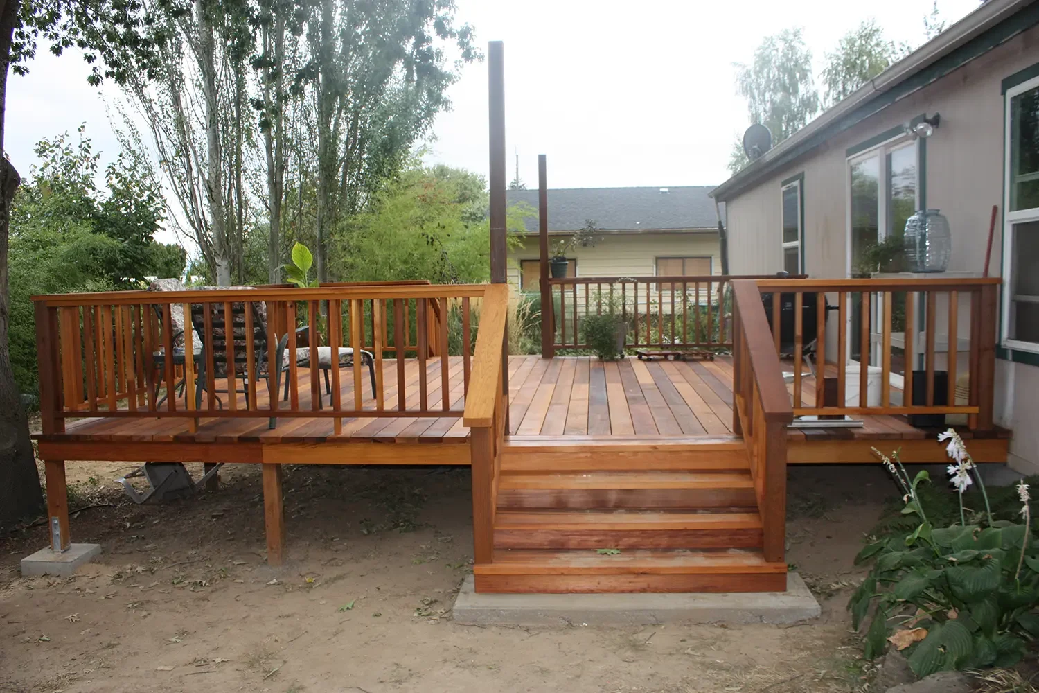 Wooden backyard deck with stairs, railing, and outdoor furniture, adjacent to a house, surrounded by trees and plants.