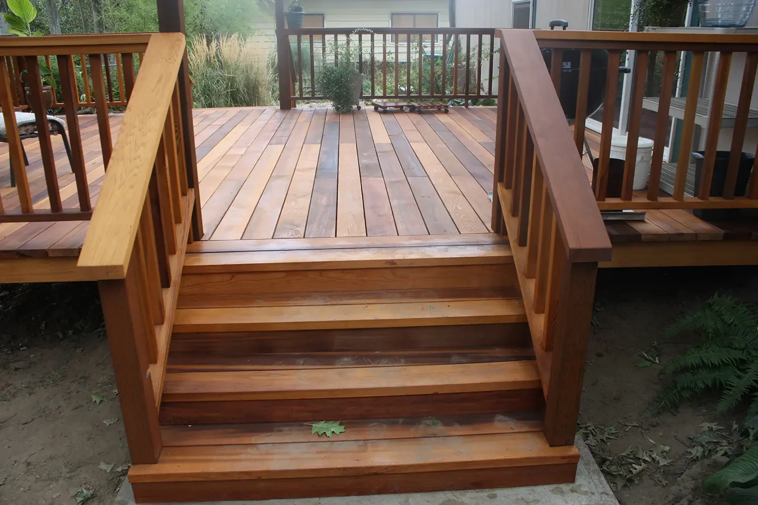 Wooden deck with stairs leading up to an outdoor patio area, surrounded by fencing and plants.