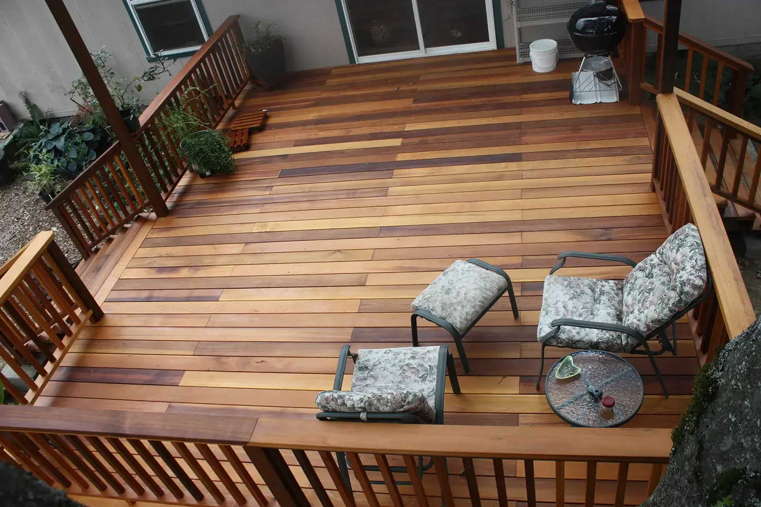 A wooden deck with outdoor furniture, including a cushioned chair, ottoman, and a glass-top table with a drink on it. There are potted plants along the railing and a grill in the corner.