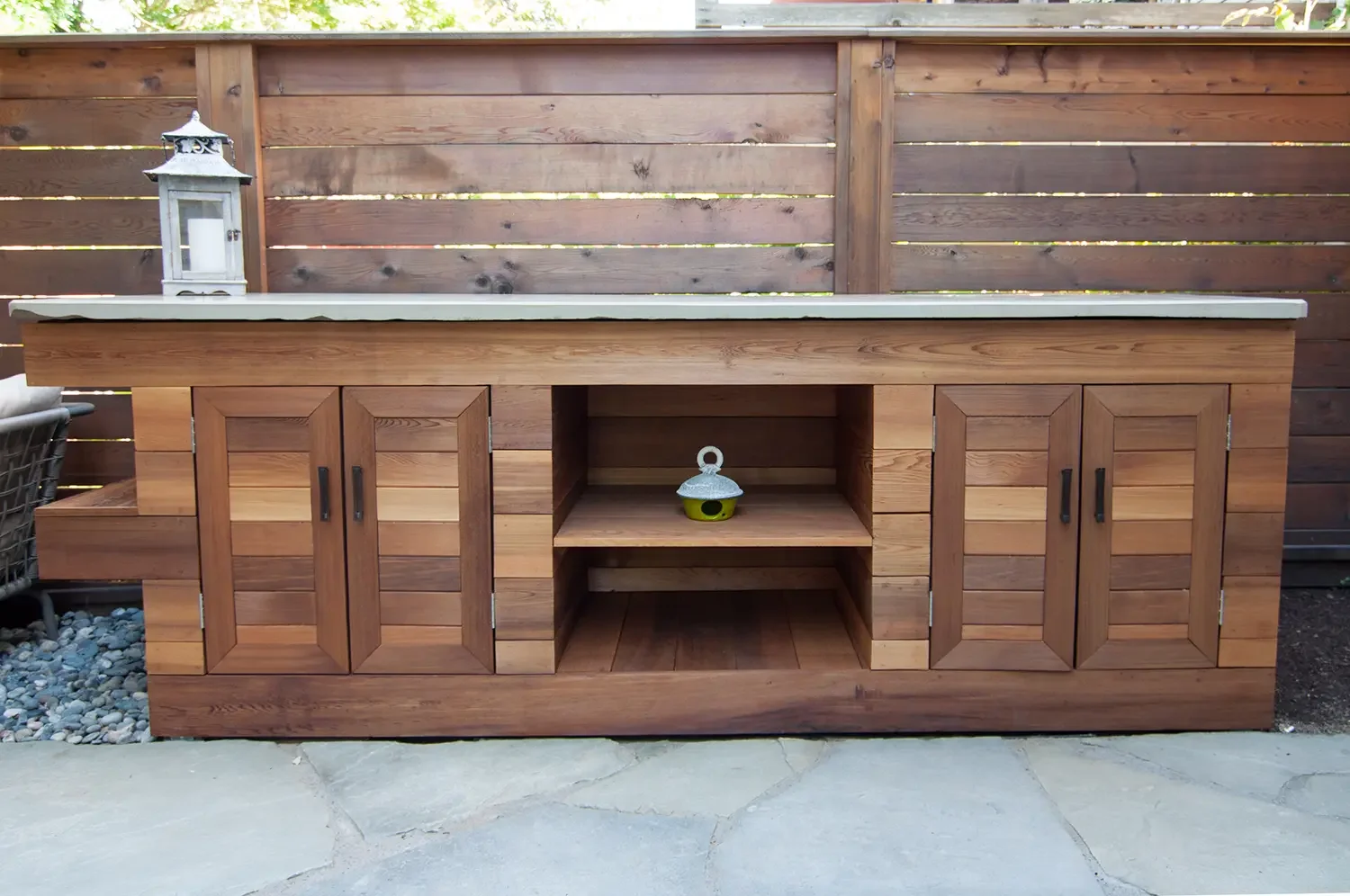 Wooden outdoor cabinet with closed doors on either side and open shelves in the center, topped with a concrete countertop, and a decorative lantern on the left side.