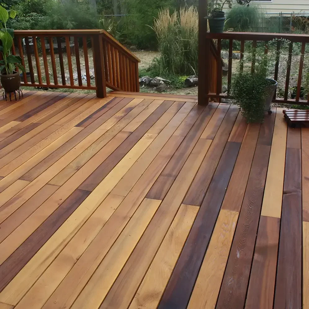 A wooden deck with orange and dark brown planks, surrounded by a wooden railing, with potted plants and a garden with grass and shrubs in the background.