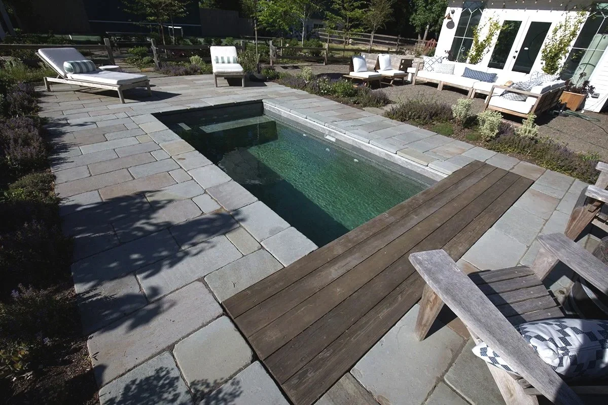 A small rectangular backyard pool surrounded by stone pavers, with outdoor seating including a lounge chair, a bench, and various chairs and sofas with cushions and pillows, on a sunny day.