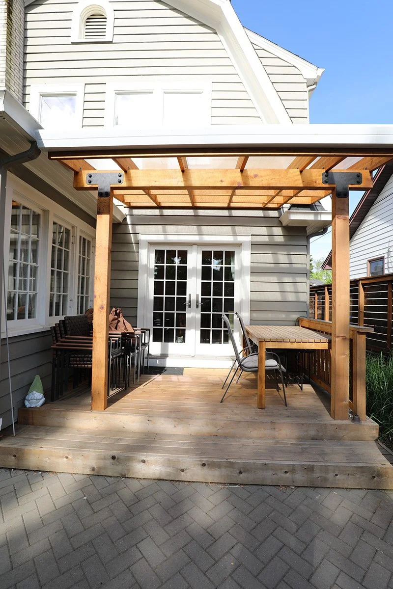 New wooden deck with a pergola attached to the back of a gray house with white trim, featuring a set of sliding glass doors, seating area, and newly laid flooring.