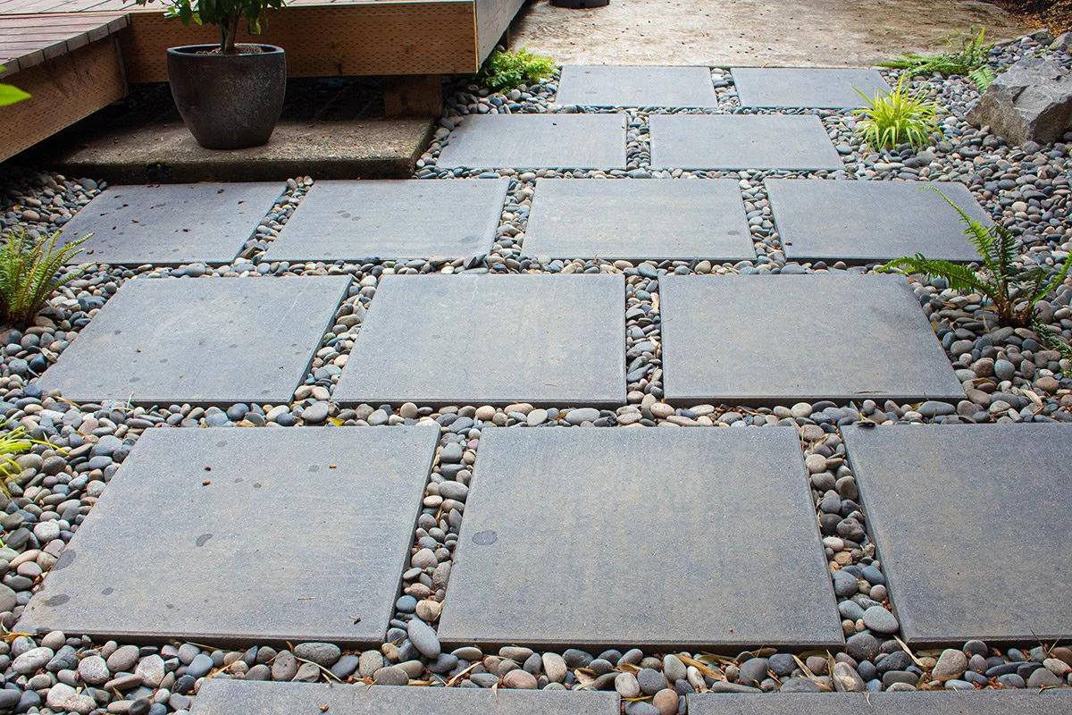 A backyard pathway made of large square gray stone pavers, bordered by small round gray and beige pebbles, with some small green plants and ferns around the sides.