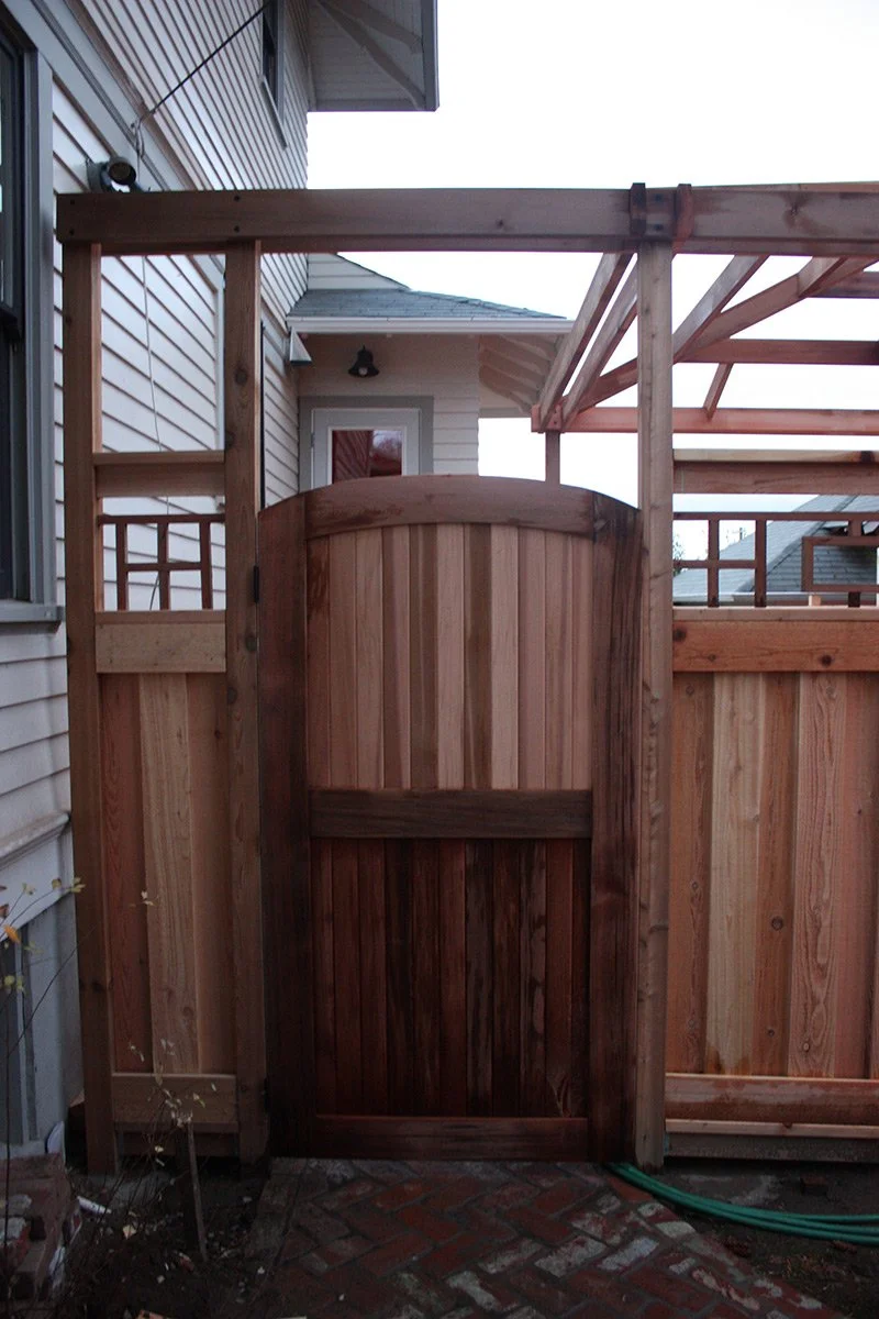New wooden gate in a backyard fence with a deck under construction on the side of a house.