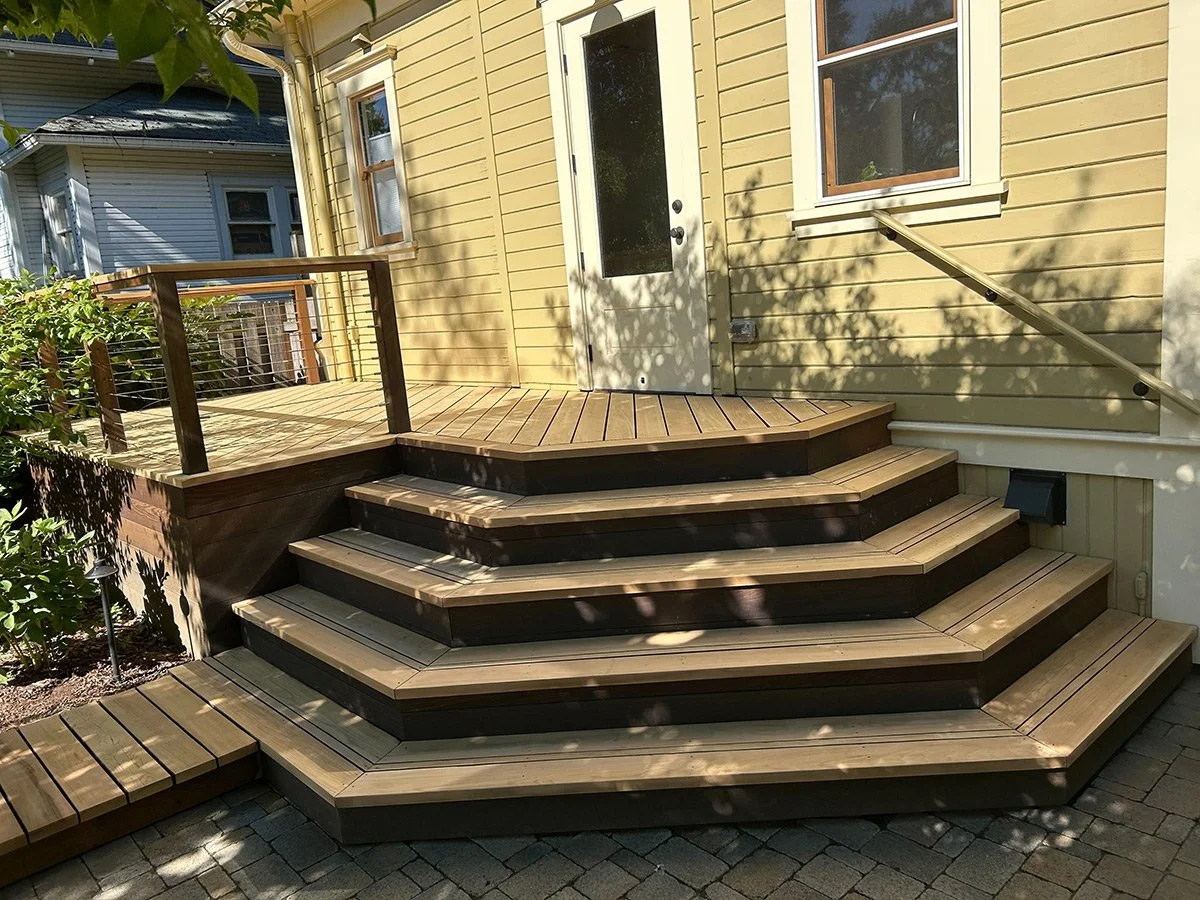 A multi-level wooden deck with stairs leading up to a door in a yellow house, with a small railing on the left side and shadows of trees cast on the house wall.