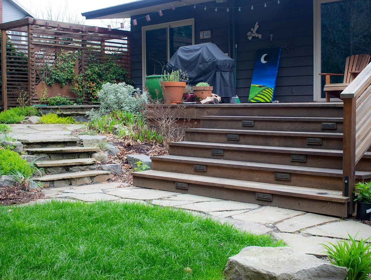 Backyard patio with stone steps leading up to a wooden deck, potted plants, a grill covered with a black cover, garden chair, and decorative outdoor art on a dark wooden house exterior.