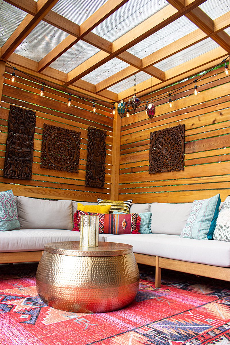 This image shows a cozy outdoor seating area with a wooden slat wall, decorative wall hangings, string lights, and a transparent roof. There is an L-shaped white sofa with colorful pillows, a metallic gold hammered coffee table, and a patterned red r
