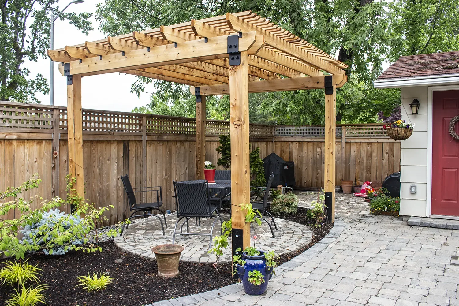 A backyard patio with a wooden pergola, outdoor dining table, and chairs, surrounded by a wooden fence, plants, and garden decorations.