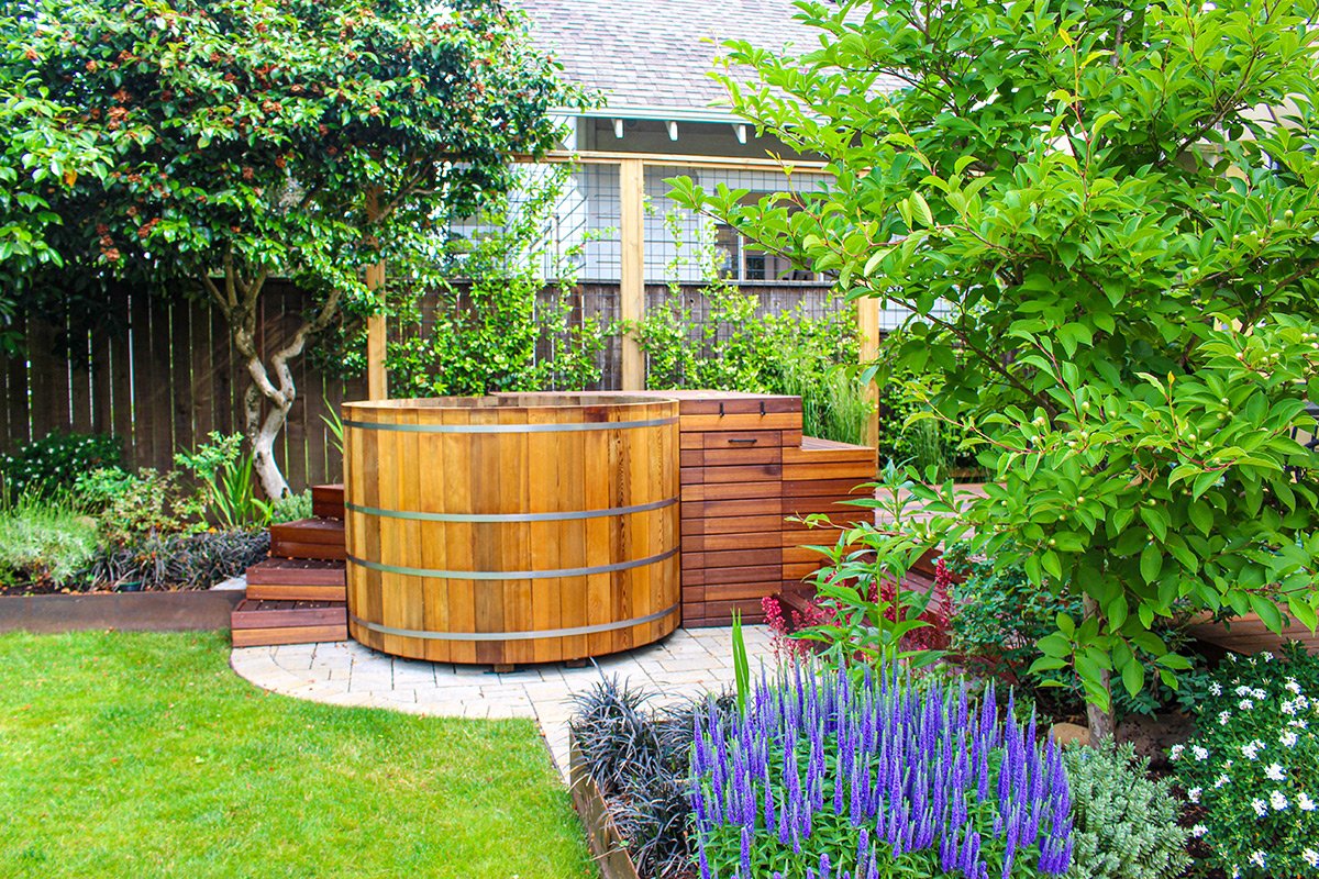Backyard garden with a hot tub made of wood, surrounded by lush green trees, shrubs, and colorful plants, on a paved patio with a wooden fence in the background.