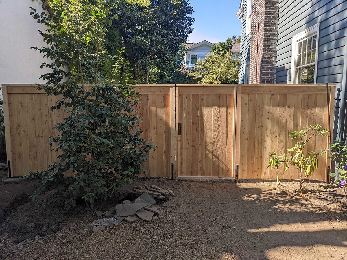 A newly installed wooden fence with a gate separates two houses in a backyard, with plants and soil in the foreground and trees and house rooftops in the background.