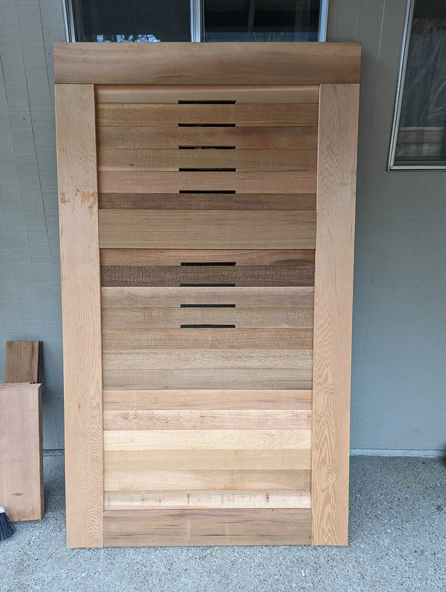 A large wooden slide composed of horizontal wooden slats with gaps in between, framed by wide vertical wooden borders, positioned outside near a building with a window.