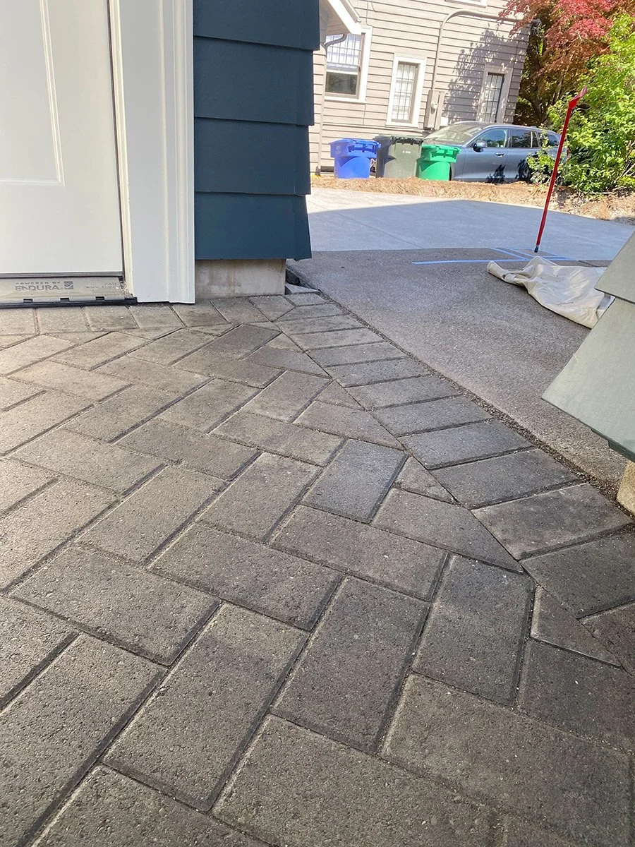 Close-up of a paved walkway transitioning from concrete to brick pavers near a house with blue siding and a white door.