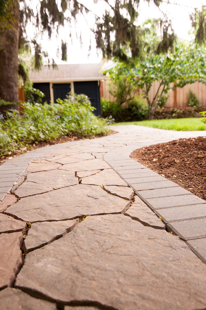 A curved stone and brick pathway in a backyard garden, with green shrubs, trees, and a wooden fence in the background.