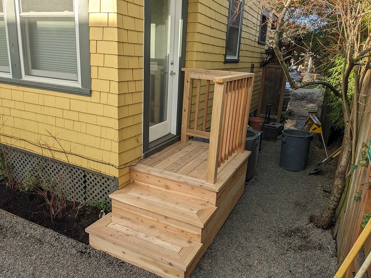 Wooden stairs leading to a small porch with a railing attached to a yellow house, with nearby plants and gardening tools.