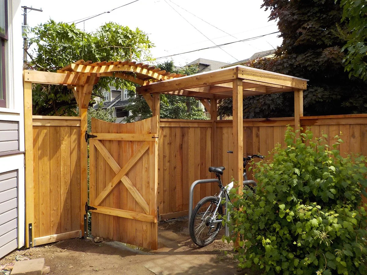 Wooden garden arbor and gate with a bicycle parked next to a tree and fenced backyard.